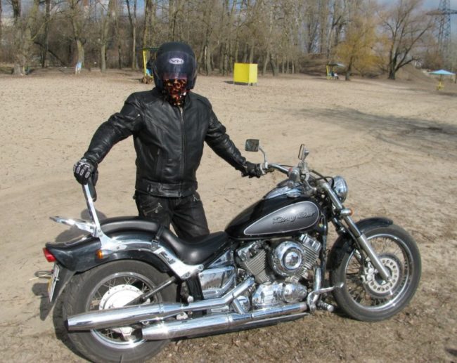 Мотоциклист в черном комбинезоне рядом с черным байком модели Yamaha Drag Star 400